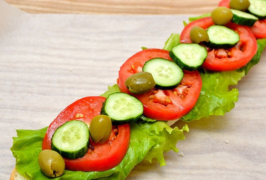 1. Этап. Бутерброды с багетом: Вторую часть багета уложите на вощеную бумагу, также тонко смажьте майонезом и уложите нарезанные овощи: лист салата, помидоры, огурец и маслины.