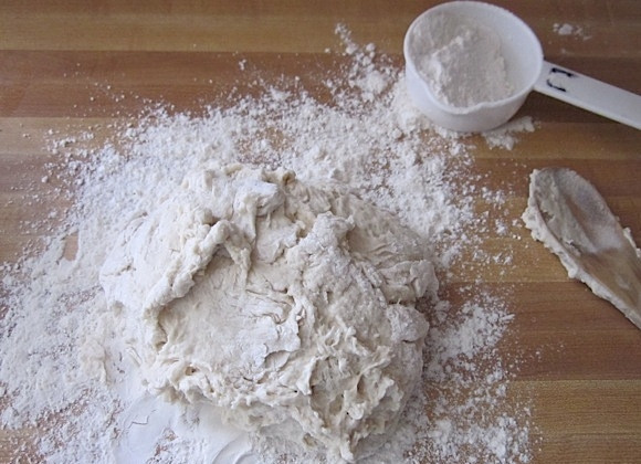 1. Этап. Вкусные бублики: 4. Добавляйте понемногу муки, чтобы тесто схватилось единым комом, затем переложите его на стол, вымешивайте тесто около 8 минут.