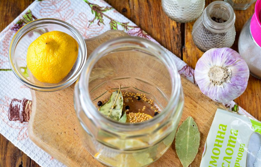 1. Этап. Огурцы с лимоном на зиму: На дно простерилизованной банки выложите специи: лавровый лист, горчицу, перец горошком, чеснок и ломтики лимона.