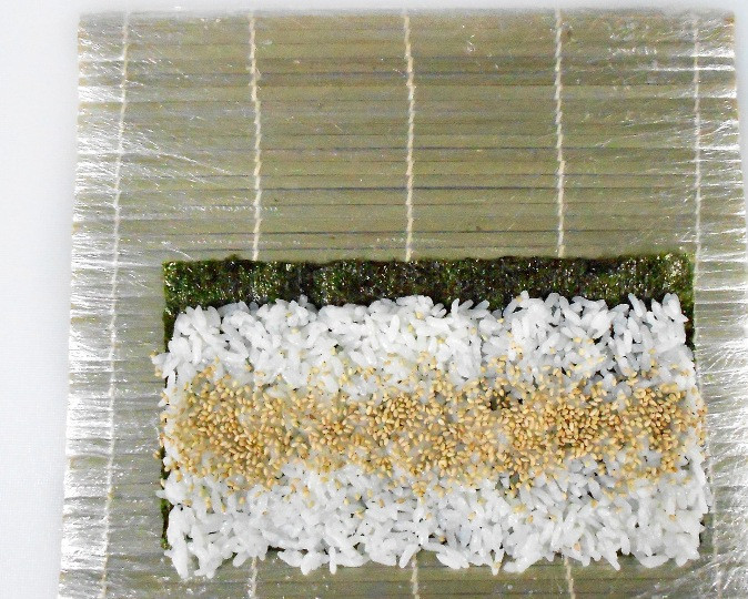 1. Этап. Роллы с угрем: Распределяем его равномерно по всему периметру, так чтобы он не доходил до одного края нории на 1 см. Теперь посередине риса делаем небольшую полоску из зерен кунжута.