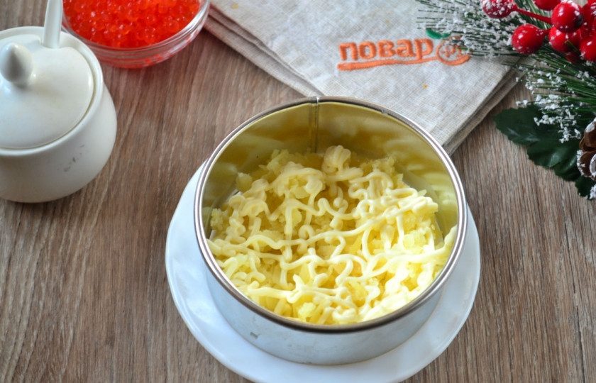 1. Этап. Сельдь под дорогой шубой: На плоскую тарелку поставьте сервировочное кольцо. На дно положите натертый на крупной терке картофель. Сверху покройте майонезной сеточкой.