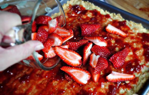 1. Этап. Клубничные пирожные с белым шоколадом: 4. Убрать форму из духовки. Нагреть клубничное варенье в микроволновой печи в течение 30 секунд, а затем смешать с лимонной цедрой. Вылить теплое варенье поверх теста. Выложить сверху нарезанную клубнику. Посыпать 1 стаканом оставшихся крошек теста.