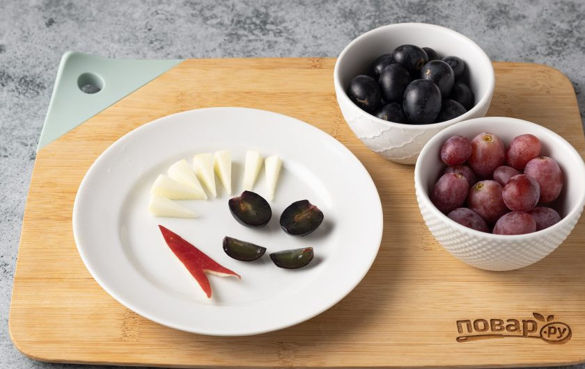 1. Этап. Фруктовая нарезка Дракон на Новый год: Из оставшегося яблока сделайте язык дракона и зубы. Еще от одной четвертинки отрежьте две дольки для глаз. Виноград помойте, обсушите и отделите от веточек. Одну черную ягоду винограда разрежьте пополам — это будут зрачки дракона, вторую разрежьте на 4 части для носа.