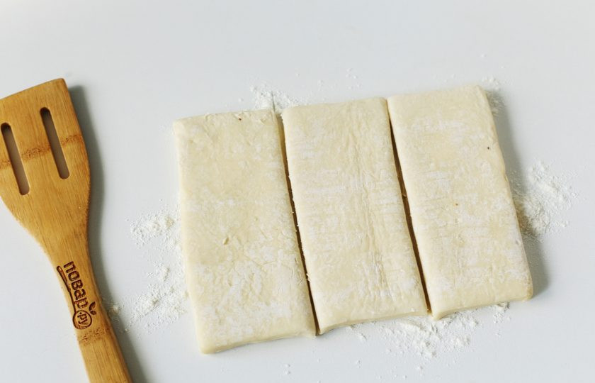 1. Этап. Слоеный пирог с мясом и грибами: Рабочую поверхность посыпьте мукой, выложите размороженный пласт теста и разрежьте его вдоль на три части как на фото.