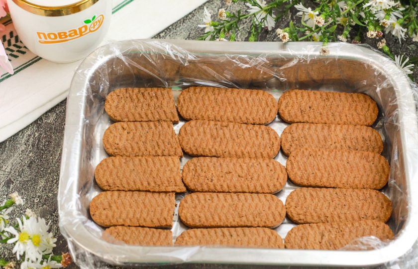 1. Этап. Торт из печенья с кремом из сметаны и сгущёнки: Застелите форму для приготовления торта пищевой пленкой и выложите нижним слоем печенье. Если требуется его поломать — разломите на части.