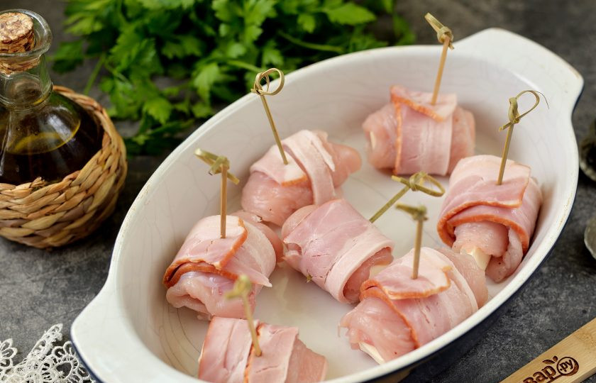 1. Этап. Куриные рулетики с плавленым сыром в беконе: Каждый рулетик заверните в ломтик бекона, скрепите заготовку шпажкой, переложите в огнеупорную форму. Отправьте форму с рулетиками в предварительно разогретую до 190 градусов духовку на 30 минут в режиме верх/низ.