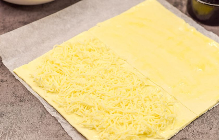 1. Этап. Слоеные палочки с сыром и беконом: Слоеное тесто раскатайте, разрежьте пополам. Куриное яйцо разбейте в миску и взболтайте вилкой. Смажьте тесто яйцом. На одну половину теста выложите тертый сыр.