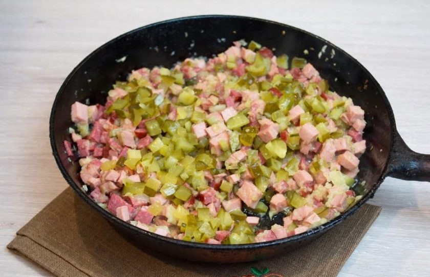 1. Этап. Солянка сборная мясная: 6. К обжаренным колбасным изделиям и луку добавьте соленые огурцы. Обжарьте все вместе 3-5 минут.