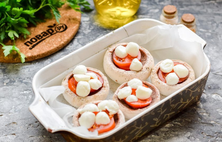 1. Этап. Шампиньоны с моцареллой в духовке: Шарики моцареллы разрежьте пополам и выложите поверх помидоров.