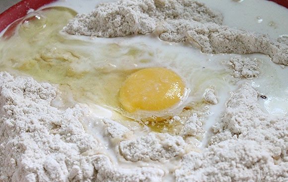 1. Этап. Простой рецепт пончиков без дрожжей: 3. В центре мучной смеси сделайте небольшое углубление, вбейте туда куриное яйцо, влейте молоко, добавьте соду, экстракт ванили, корицу, мускатный орех, сахарную пудру.