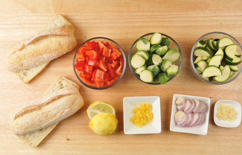 1. Этап. Багет с запечёнными овощами: 1. Подготовьте ингредиенты. Измельчите чеснок. С лимона натрите цедру. Лук нарежьте кольцами, перец - кубиками. Багет разделите пополам. Брюссельскую капусту нарубите половинками, а цукини - полукружками.