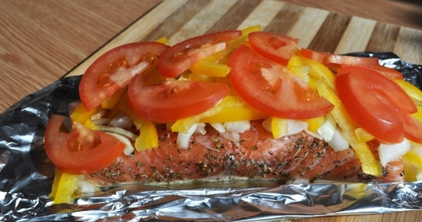 1. Этап. Семга, запеченная в фольге: Каждый кусок рыбы выкладывайте на отдельную фольгу, чтобы её можно было завернуть вдвое. Сверху уложите слой лука, перца, а наверх — помидор. Всё полейте оливковым маслом.