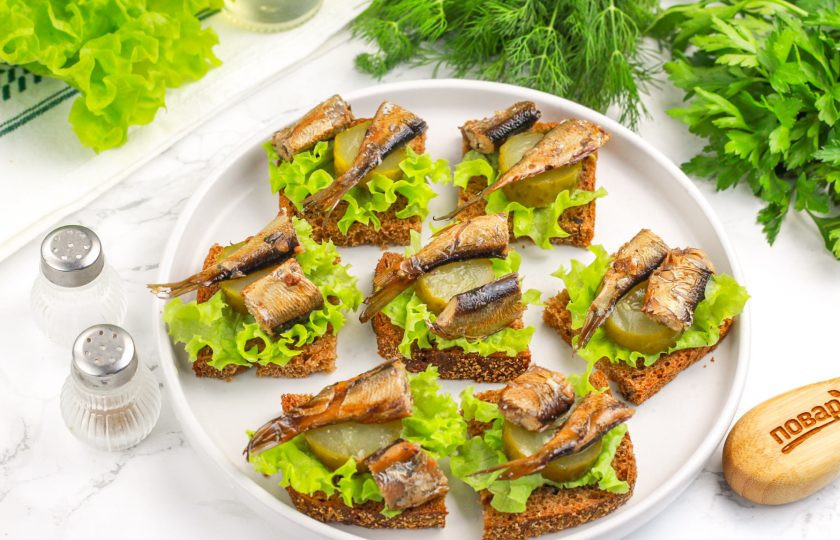 1. Этап. Канапе со шпротой: Каждую рыбку стряхните от излишков масла и разрежьте пополам. Выложите на соленый огурец. Украсьте канапе зеленью укропа или петрушки.