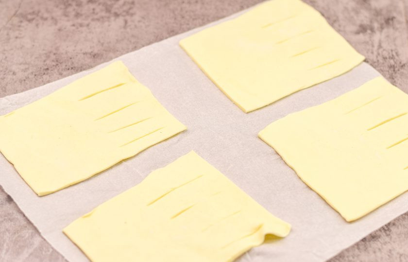 1. Этап. Слоеные пирожки с рикоттой и шпинатом: Слоеное тесто раскатайте немного, разрежьте на прямоугольники. Половину каждой заготовки разделите визуально пополам, на одной половине сделайте разрезы, как показано на фото.