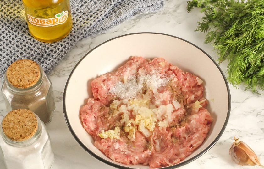 1. Этап. Колбаски «По-балкански» с салатом из запеченных овощей: Мясной фарш выложите в глубокую емкость. Четверть очищенного репчатого лука и зубчик чеснока натрите на терке с мелкими ячейками. Выложите массу в емкость с фаршем, всыпьте 3 щепотки соли и молотый черный перец.