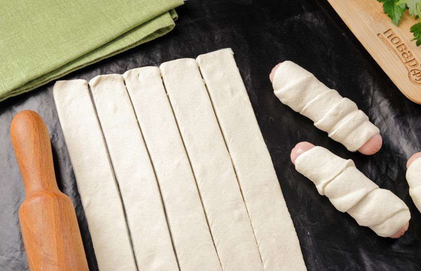 1. Этап. Сосиски в слоеном бездрожжевом тесте в духовке: Оберните каждую сосиску полоской слоёного теста. Если полоски получились длинными, то можете обматывать внахлест.