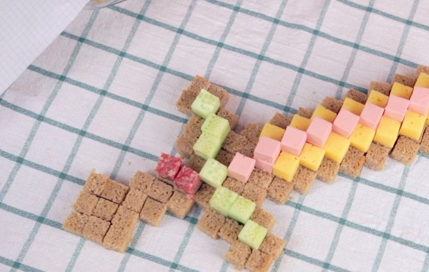 1. Этап. Съедобный золотой меч из Майнкрафта: 3. Далее на блюде, на котором будете подавать меч, выложите сначала основу из хлеба, потом положите полоски из сыра и колбасы, затем рукоятку меча выложите из огурца и копченой колбасы.