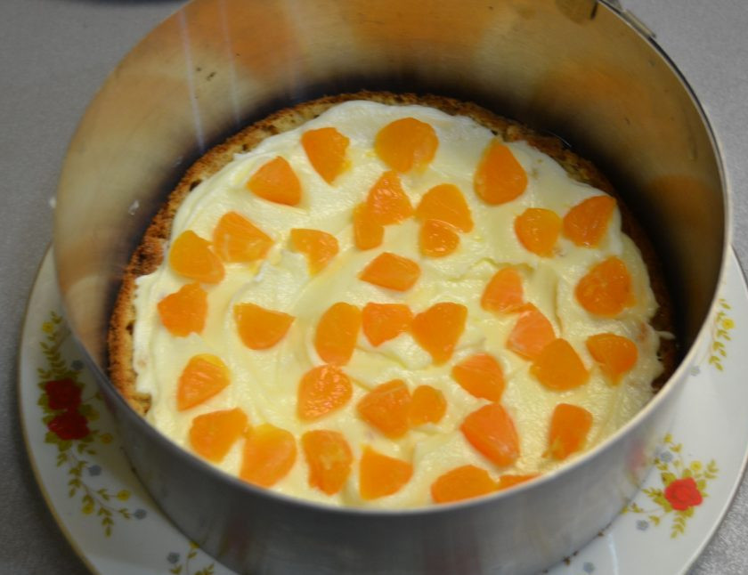 1. Этап. Мандариновый торт на Новый год: Собирайте торт в кольце, уложите на низ первый корж, выложите 1/4 часть крема, на крем выложите кусочки мандарина.