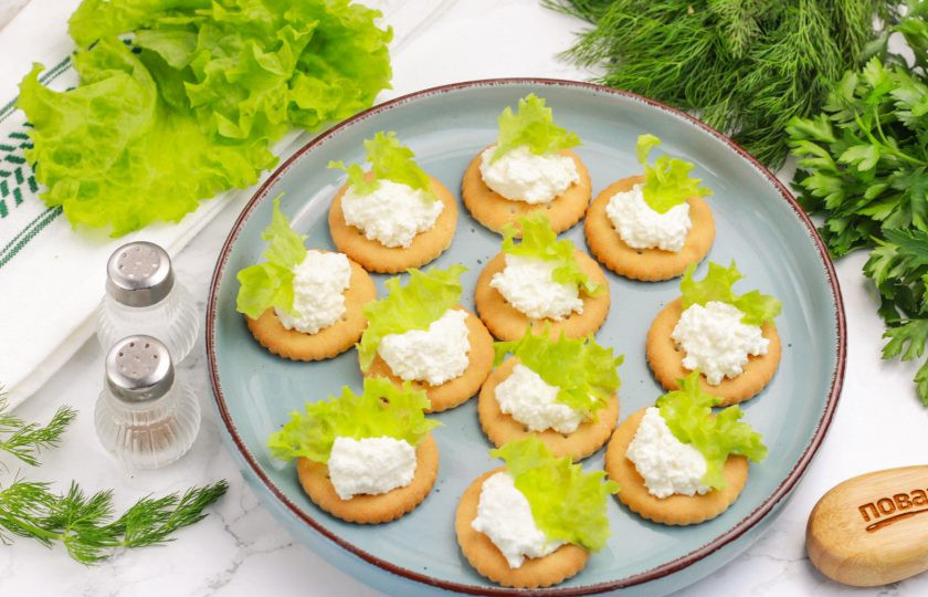 1. Этап. Канапе на крекерах на праздничный стол: Промойте листья салата и нарвите небольшие полосочки. Выложите их на творожную массу.