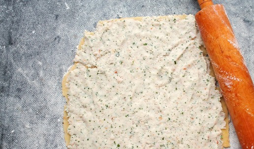 1. Этап. Закусочное печенье с ветчиной и сыром: Рабочую поверхность немного присыпьте мукой и раскатайте в прямоугольник 25 на 35 сантиметров и толщиной 2-3 миллиметра. Нанесите сверху начинку, прижимая. Сверните все в плотный рулет, заверните в пленку и уберите в морозильную камеру на 30 минут. Это позволит нарезать рулет ровно и нужной толщины.