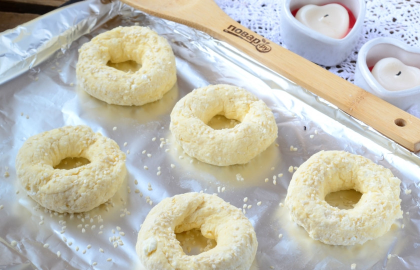 1. Этап. Творожные бублики с сыром: Полученное тесто разделите на 5 равных частей. Можно сделать и больше бубликов, но тогда они получатся более тонкими и не такими нежными. Из каждой части сделайте колбаску толщиной в указательный палец, а затем соедините, чтобы образовалось колечко. Выложите колечки на противень, присыпьте сверху кунжутными семечками.