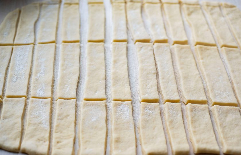 1. Этап. Хворост в духовке на кефире: Рабочую поверхность припылите мукой и раскатайте тесто в пласт толщиной примерно в 1-2 мм. Нарежьте пласт теста на полосы шириной в 2-3 см. Затем каждую полосу нарежьте в форме прямоугольника.