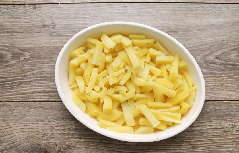 1. Этап. Картофель с беконом в сметанной заливке: Картофель очистите и порежьте крупной соломкой. Отварите в соленой воде до полуготовности. Спустя время откиньте на сито, чтобы слить отвар. Форму для выпечки смажьте растительным маслом и переложите в нее картофель.