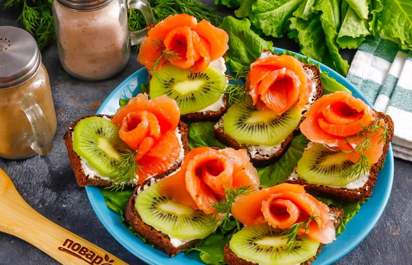 1. Этап. Бутерброды с красной рыбой и киви: Нарезку слабосоленой красной рыбы нарежьте лентами примерно в 1,5-2 см. шириной. Сверните их в виде роз и поместите на другой край бутербродов.