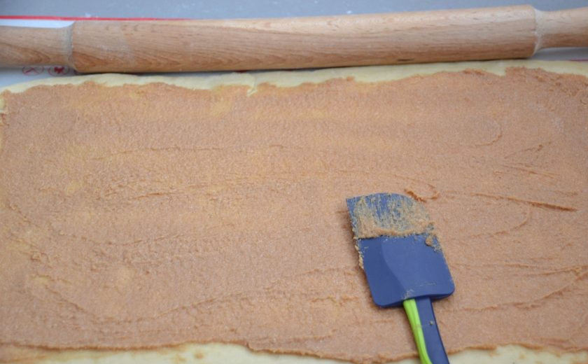 1. Этап. Финские булочки с корицей и кардамоном: Присыпьте рабочую поверхность стола мукой, раскатайте тесто в прямоугольный пласт, равномерно нанесите на него начинку, не доходя до края примерно 1,5-2 см.