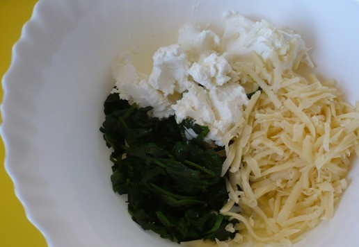 1. Этап. Рождественская елка из слоеного теста: Выложите приготовленный шпинат на разделочную доску, нарежьте кусочками и переложите в миску, добавьте творожный сыр и натертый на крупной терке полутвердый сыр. Затем перемешайте до получения однородной массы.