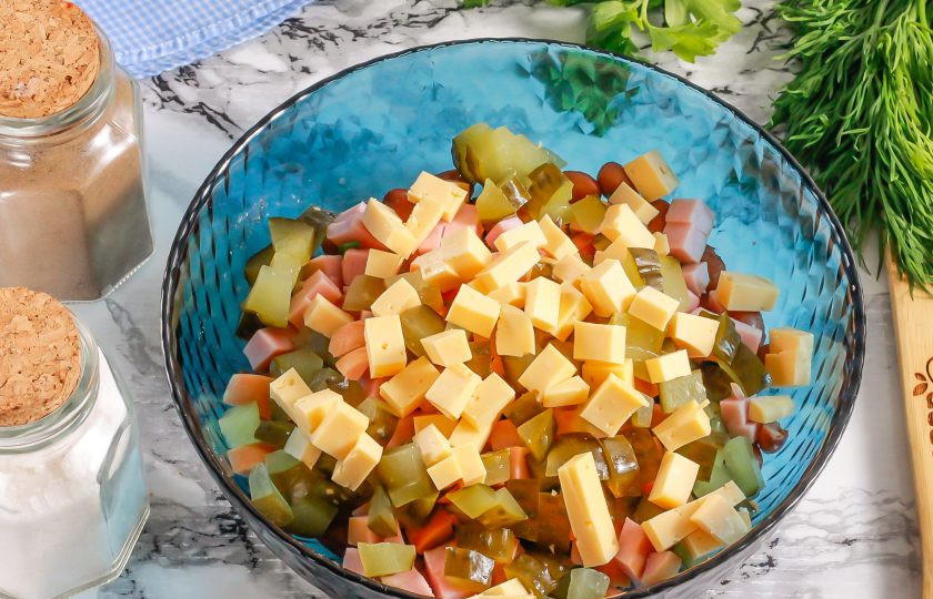 1. Этап. Салат с фасолью, сосисками, сыром и сухариками: Твердый сыр нарежьте такими же кубиками и соедините с другими нарезками в емкости. С соленого огурца срежьте хвостики и нарежьте его кубиками, добавьте к сыру.