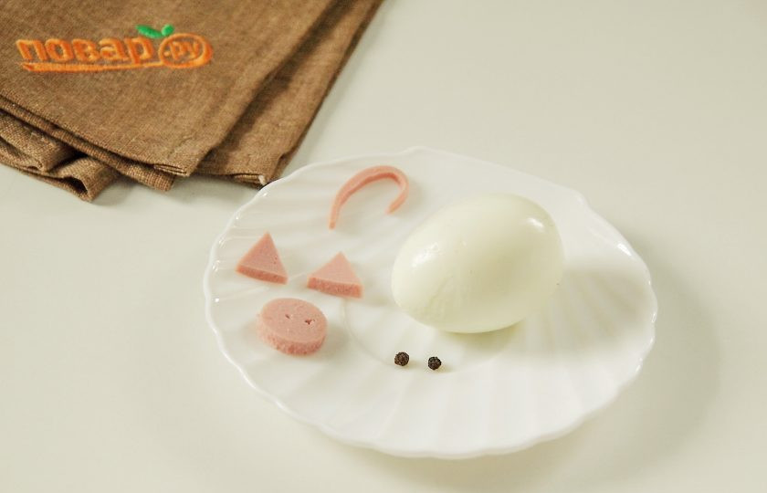 1. Этап. Слоеный салат Новогодний: Так как салат предлагаю подать к новогоднему столу, оформим его соответственно - украсим свинкой из вареного яйца. Из кусочка колбаски вырежем ушки, пятачок и хвостик. Горошины перца послужат глазами.