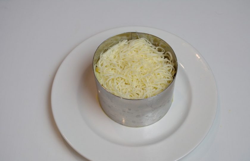 1. Этап. Салат 8 Марта с кальмарами: При помощи сервировочного кольца оформите салат. Выложите салат в кольцо. Утрамбуйте его. Натрите на терке твердый сыр. Сделайте 