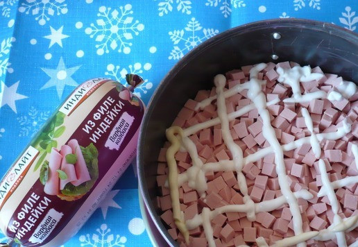 1. Этап. Салат к Новогоднему столу: Салат я выкладывала слоями в разъемную форму для запекания (d-18 см). Установить её на плоское блюдо. Первым слоем выложить нарезанную вареную колбасу и сделать сеточку из майонеза.