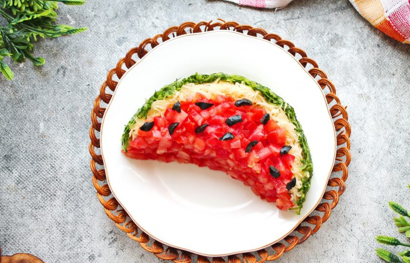 1. Этап. Салат Арбузная долька с курицей и грибами: У огурца натрите на крупной терке только зеленую часть, все остальное не понадобится. Отожмите зеленую массу и сформируйте зеленую корку, прижав к сырному слою. Маслины нарежьте на кусочки и разложите на помидорный слой как арбузные косточки.