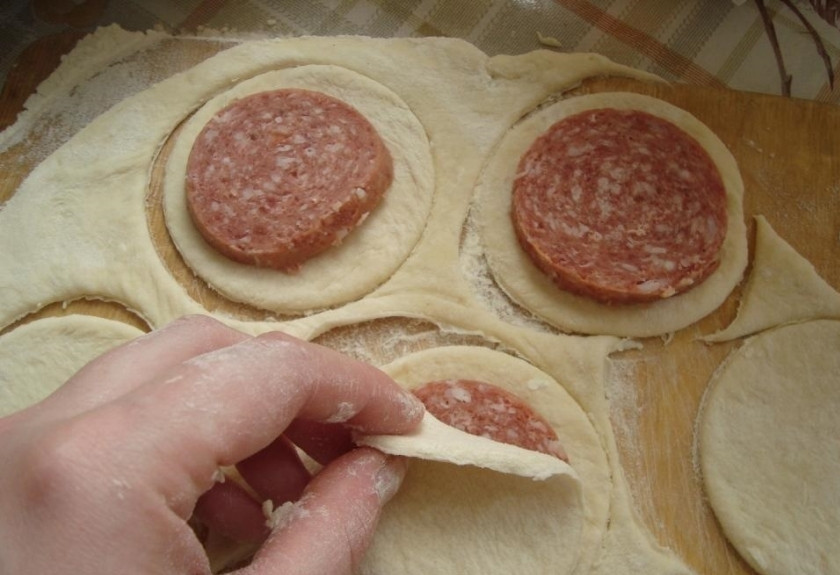 1. Этап. Колбаса в тесте: 2. От общей массы отщипываем кусочек, раскатываем его толщиной 2-3 миллиметра, чтобы было тонко, но не рвалось. Стаканом вырезаем кружочки. Колбасу нарезаем кусочками (3-5 миллиметра) и выкладываем ее внутрь. Вторым кружочком теста накрываем и хорошенько защипываем колбасу внутри.