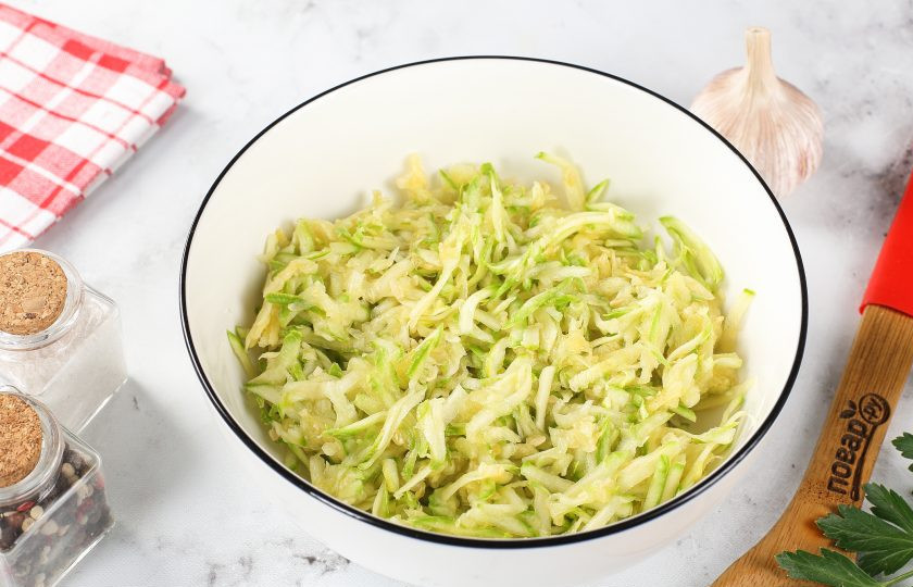 1. Этап. Запеканка из кабачков с копчёной колбасой и сыром: Кабачок натрите на крупной тёрке, добавьте щепотку соли, перемешайте и оставьте на 10 минут. Затем отожмите от выделившегося сока и переложите в глубокую миску.