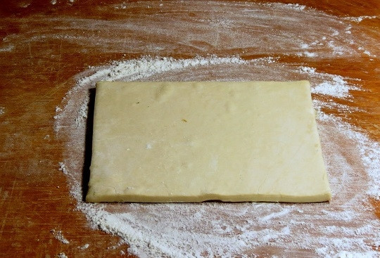 1. Этап. Выпечка из слоеного теста в духовке: 2. Горсть муки высыпьте на рабочую поверхность, выложите тесто. Оно должно быть не слишком замороженным, чтобы с ним можно было легко работать. Раскатывать его вы можете как очень тонко, так и совсем немного, от этого будет зависеть толщины коржа. Ориентируйтесь также на размер вашей формы для запекания.