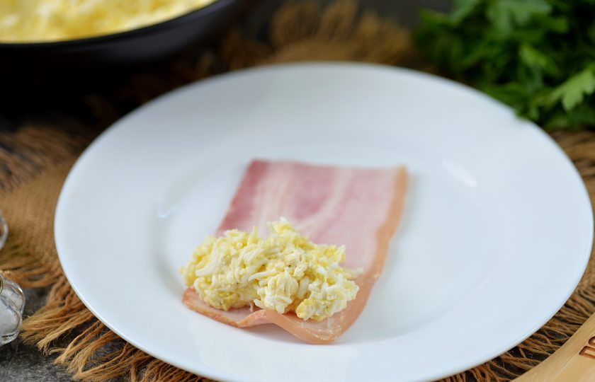 1. Этап. Рулетики из бекона с сыром и яйцом: Если полоски бекона слишком длинные, разрежьте их пополам. Выложите на каждый ломтик бекона по чайной ложке начинки из яиц и сыра.