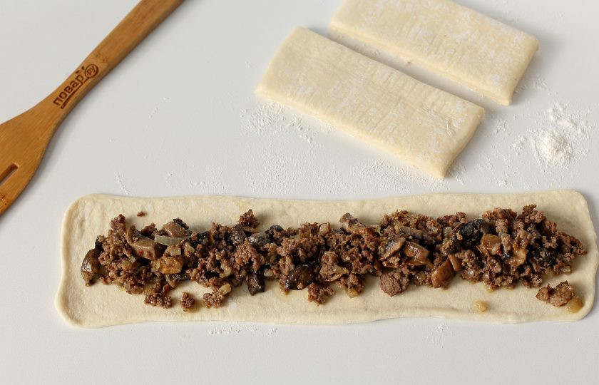 1. Этап. Слоеный пирог с мясом и грибами: Каждую полоску теста раскатайте на узкие длинные ленты шириной 10 см. Выложите в середину каждой длинной ленты мясной фарш с грибами.