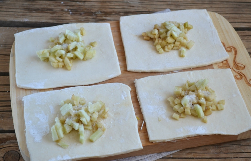 1. Этап. Конвертики из слоеного теста с начинкой: Тесто порежьте на квадраты примерно 6 на 6 см. Выложите на них яблоки, присыпьте сахаром. Скрепите уголки квадрата, сформируйте конвертик. Выложите конвертики на противень швом вниз. Смажьте яйцом.
