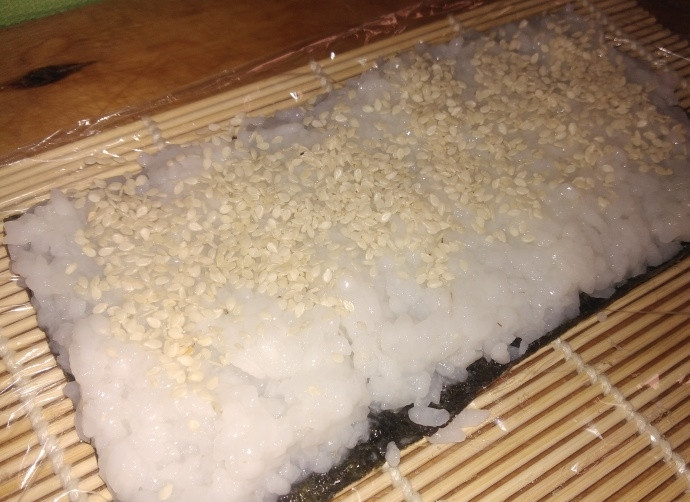 1. Этап. Роллы Аляска: 3. Бамбуковый коврик застелите пищевой пленкой, выложите лист нори и смочите его слегка водой, распределите рис, оставляя 1 сантиметр с одного края. Посыпьте рис кунжутом.