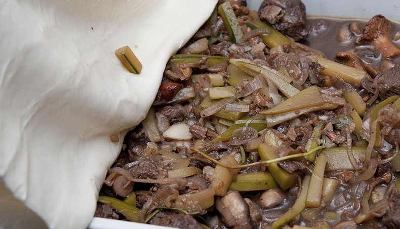 1. Этап. Мясной пирог под слоеным тестом: Когда мясо будет готово, снимаем сковороду с огня, а все ее содержимое перекладываем в жаропрочную форму для запекания. Сверху накрываем начинку раскатанным слоеным тестом, заворачивая края вовнутрь.