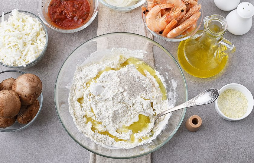 1. Этап. Пицца с креветками и грибами: В глубокой миске смешайте муку с дрожжами и солью. Добавьте 2 столовые ложки оливкового масла и начинайте замешивать тесто, постепенно подливая теплую воду. Количество воды зависит от сухости муки и может колебаться. Замесите мягкое тесто, отстающее от рук и от миски. В процессе вымешивания используйте оставшуюся ложку масла.