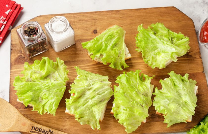 1. Этап. Праздничные бутерброды Змейка: Смажьте нижнюю часть бутербродов майонезом и кетчупом или любым другим соусом по вкусу. Накройте листиком салата.