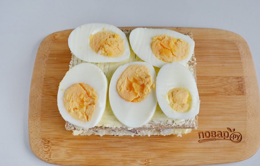 1. Этап. Закусочный бутербродный торт с рыбой: Накройте селедку кусочком серого хлеба, смажьте его маслом и положите сверху колечки вареного яйца.