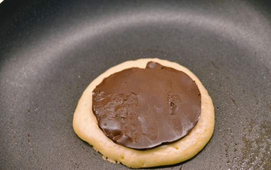 1. Этап. Панкейки с начинкой внутри: Из шоколадной пасты формируем кругляши, намазываем их на пергамент. Отправляем на полчасика в морозилку. После на разогретую сковородку наливаем масло, выкладываем две столовые ложки нашего теста, а сверху - замороженный кружочек шоколадной пасты.