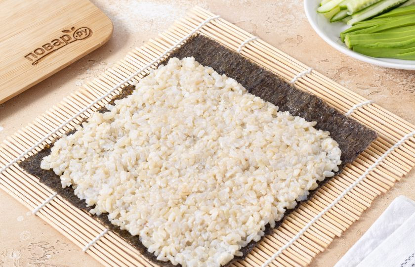 1. Этап. Роллы с бурым рисом: Положите на бамбуковый коврик лист нори гладкой стороной вниз. Мокрыми руками выложите бурый рис ровным слоем, оставив свободными пару сантиметров с одного края.