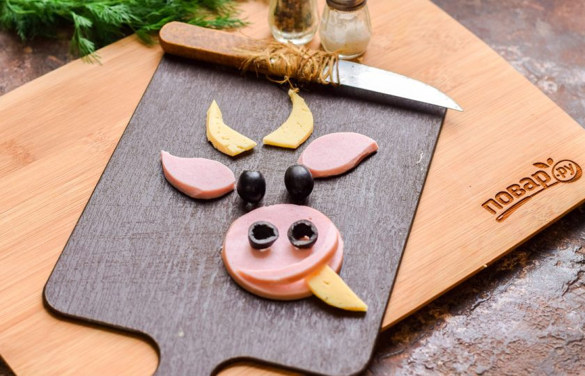 1. Этап. Слоеный салат с курицей Бычок: Из колбасы, сыра и маслин вырежьте заготовки для лица быка. Колбаска хорошо подходит вареная.