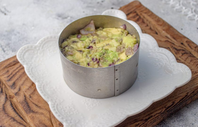 1. Этап. Праздничный легкий салат с семгой и авокадо: В сервировочное кольцо диаметром 8-10 см. установите на тарелку. Первым слом выложите авокадо с луком и плотно утрамбуйте.